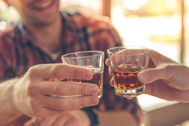 Zwei Personen stoßen mit Whiskygläsern an, in einer gemütlichen, hellen Umgebung.