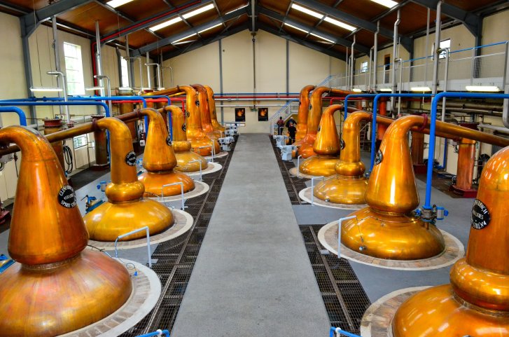 Kupferne Destillierapparate stehen still in einer gut beleuchteten Brennereihalle mit hohen Decken.