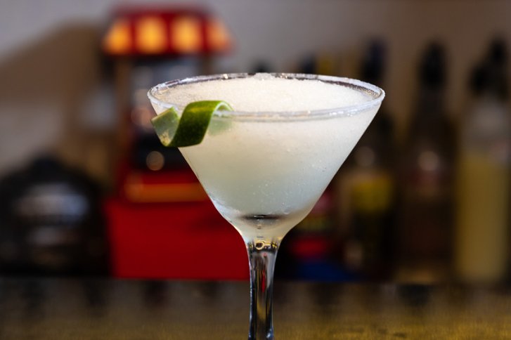 Cocktailglas mit Crushed Ice und Limettenscheibe steht auf einer Bar vor unscharfem Hintergrund.