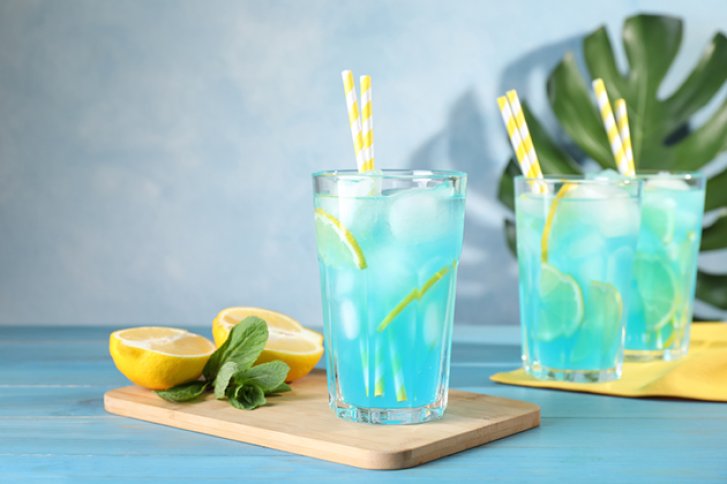 Drei blaue Cocktails mit Zitronenscheiben und Strohhalmen stehen auf blauem Tisch, umgeben von Zitronen.