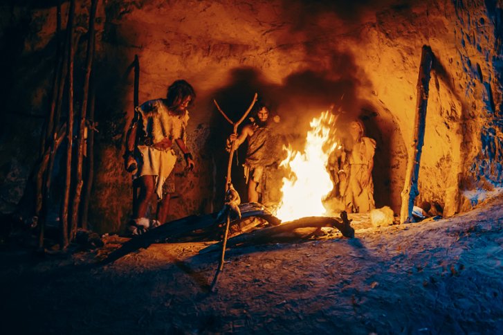 Höhlenmenschen stehen um ein loderndes Feuer in einer felsigen Höhle mit Stöcken.