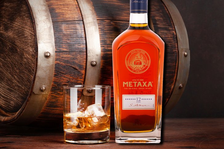 Flasche Metaxa steht neben gefülltem Glas, vor einem hölzernen Fass in rustikalem Ambiente.