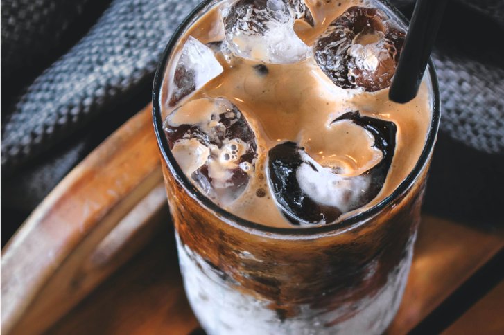 Ein Glas Eiskaffee mit Eiswürfeln steht auf einem Holztisch, schwarzer Strohhalm eingefügt.