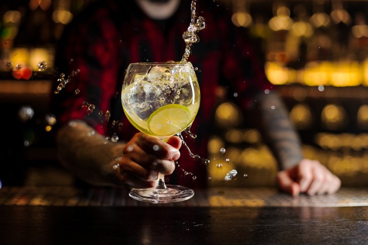 Ein Glas mit spritzendem Getränk wird von tätowierter Hand in einer Bar gehalten.