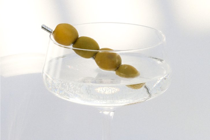 Ein Martini-Glas hält vier Oliven auf einem Spieß in klarem Getränk, minimalistischer Hintergrund.