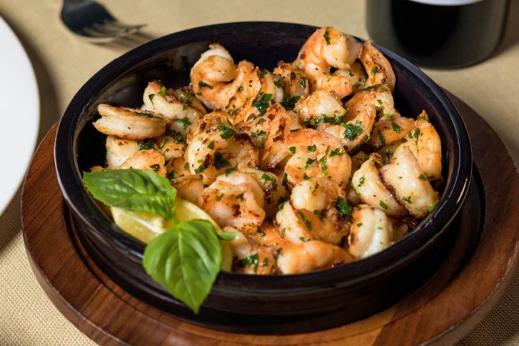 Garnelen liegen gewürzt in einer schwarzen Schüssel auf einem Holzteller, dekoriert mit Basilikum.