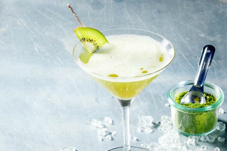 Ein grüner Cocktail mit Kiwi-Deko steht neben Glas mit grünem Pulver auf Eis.
