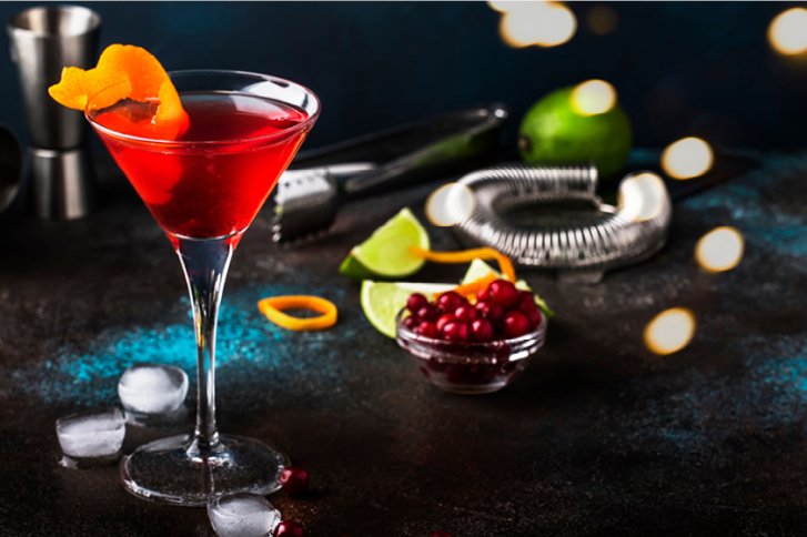 Roter Cocktail in Martini-Glas, dekoriert mit Orangenschale, umgeben von Limetten und Beeren.