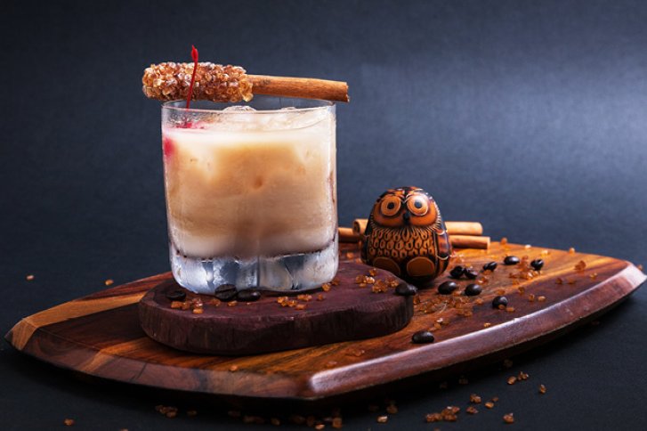 Ein cremiger Cocktail mit Zimt und Kandiszucker auf Holzbrett, neben Deko-Eule und Kaffeebohnen.