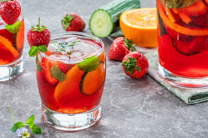 Ein Glas roter Cocktail mit Eis, Erdbeeren und Orangenscheiben auf grauem Untergrund.
