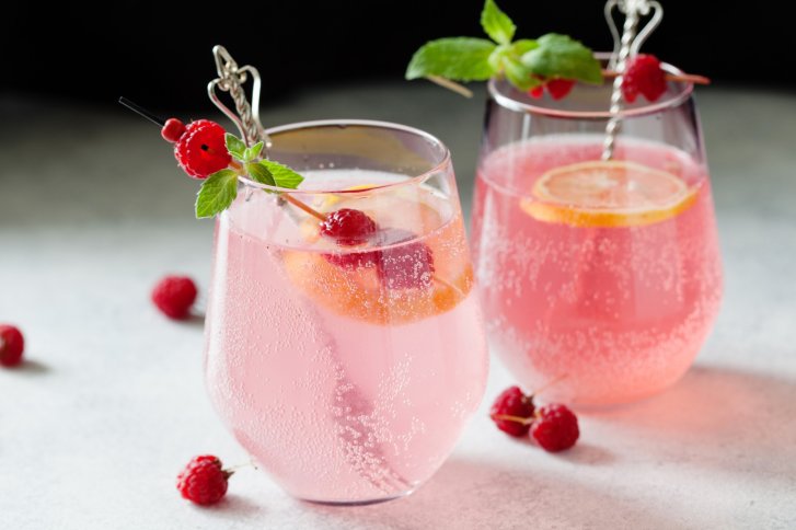 Zwei rosa Cocktails mit Zitronenscheiben und Himbeeren, in Gläsern mit Herzspießen dekoriert.
