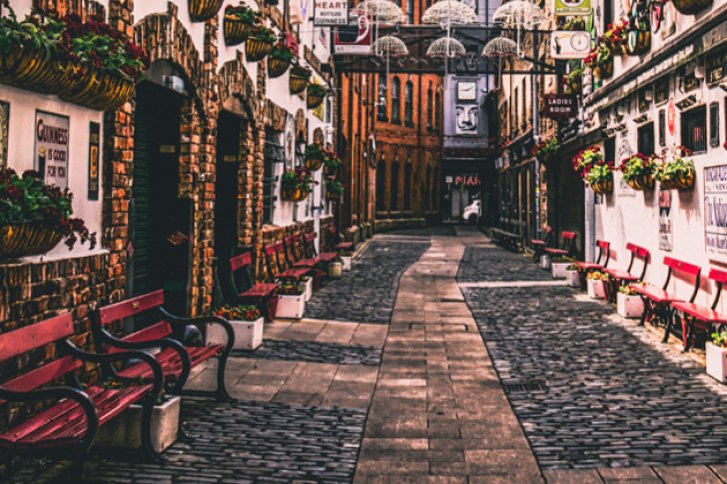 Gepflasterte Gasse mit roten Bänken; hängende Blumen und Regenschirme; alte Gebäude umgeben.