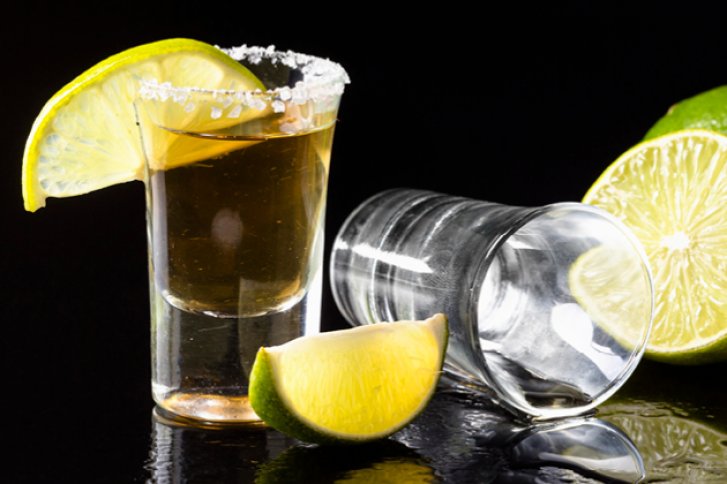 Ein Tequilaglas mit Salzrand steht, daneben Limettenscheiben, auf schwarzem, glänzendem Untergrund.