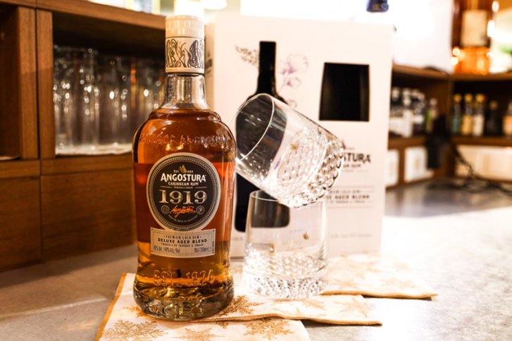 Rumflasche steht neben zwei Gläsern auf Theke, vor Regal mit Trinkgläsern.