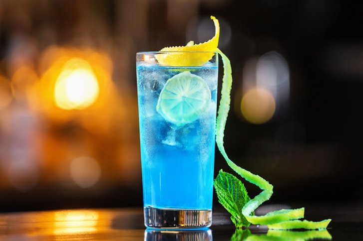 Blaues Getränk im Glas mit Limettenscheibe, Zitronenschale und Minze, auf einem Tresen.