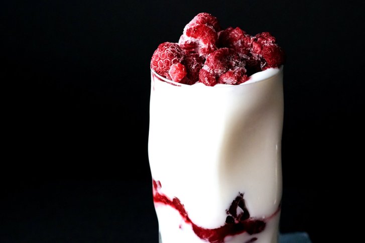 Ein Glas gefüllt mit Joghurt und gefrorenen Himbeeren vor schwarzem Hintergrund.