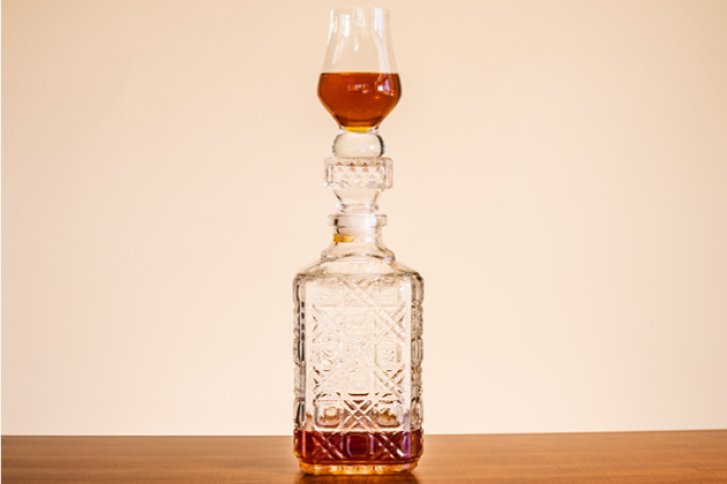 Karaffe mit Muster steht, Glas obenauf, gefüllt mit braunem Getränk, vor beigem Hintergrund.