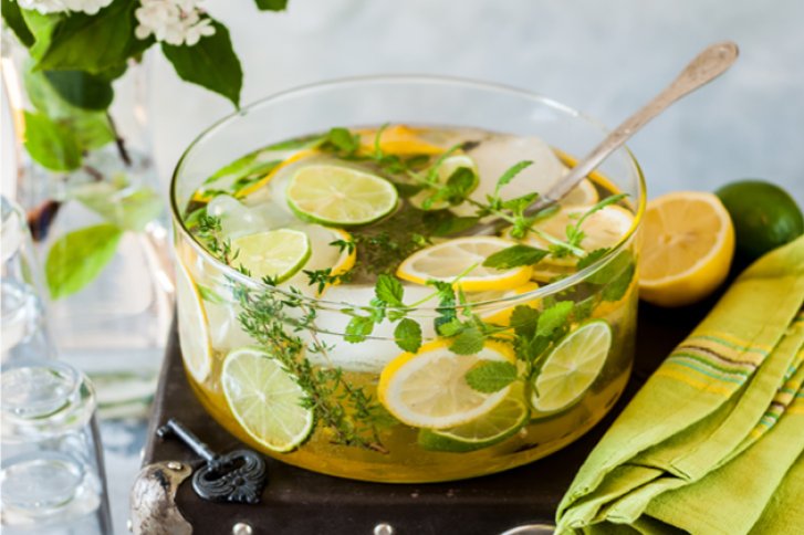 Eine große Glasschale mit Kräutern, Zitronenscheiben und Limetten steht auf einem gedeckten Tisch.