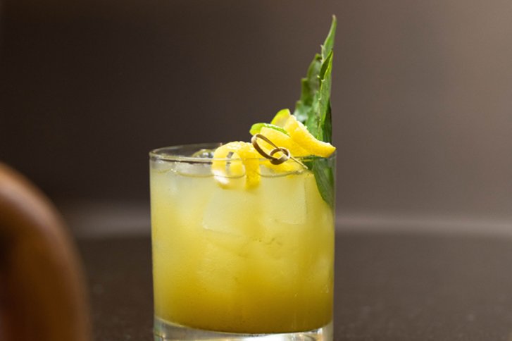 Ein Glas mit gelbem Cocktail, dekoriert mit Zitrone und Ananasblatt, steht auf Tisch.