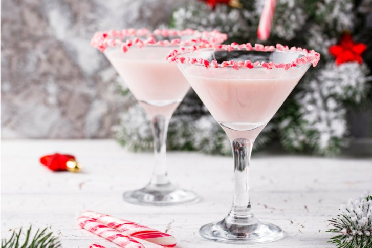 Zwei Cocktailgläser mit rosa Flüssigkeit und Zuckerrand stehen auf einem weißen, weihnachtlich dekorierten Tisch.