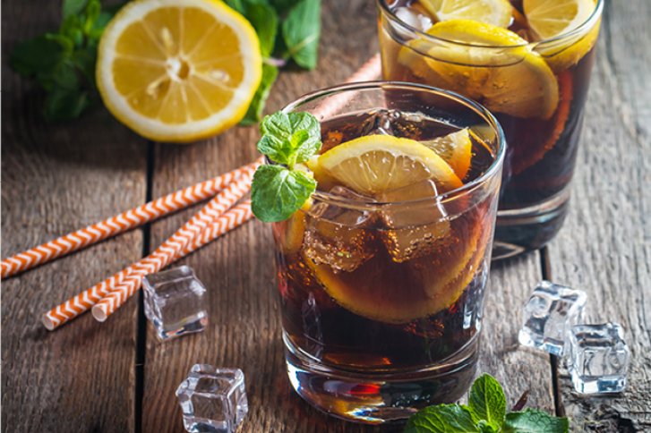 Gläser mit Cola, Zitrone und Minze stehen auf einem Holztisch mit Eiswürfeln und Strohhalmen.