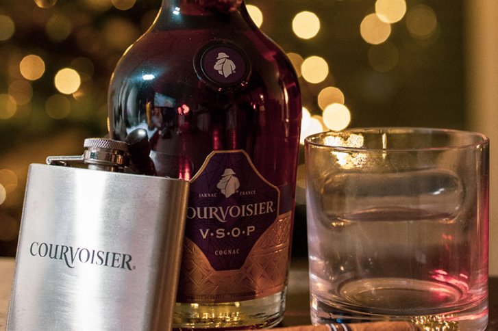 Flasche Cognac, Taschenflasche und Glas auf Tisch, umgeben von unscharfen Lichtern im Hintergrund.