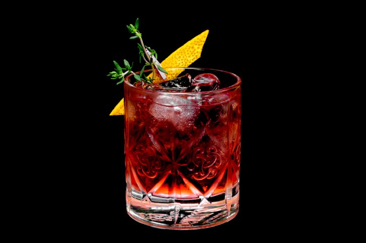 Kristallglas enthält roten Drink mit Eiswürfeln, dekoriert mit Thymian und Zitronenschale, vor schwarzem Hintergrund.