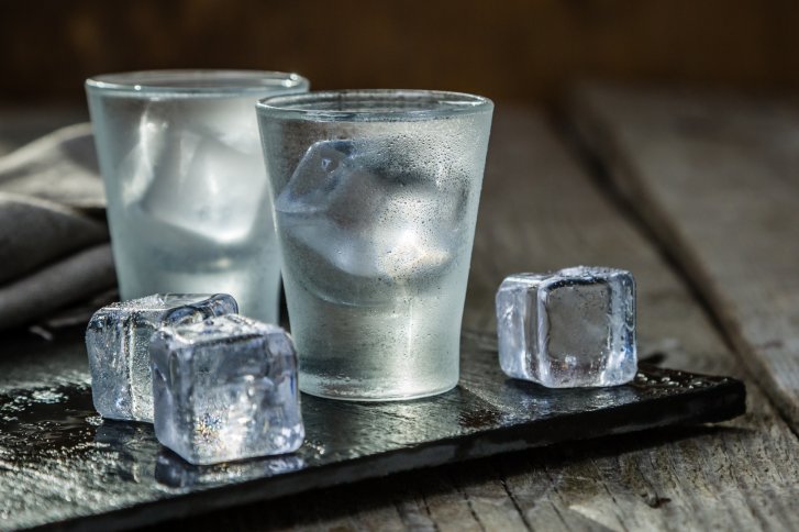 Zwei Gläser mit Eiswürfeln stehen auf einem nassen, dunklen Untergrund aus Stein.