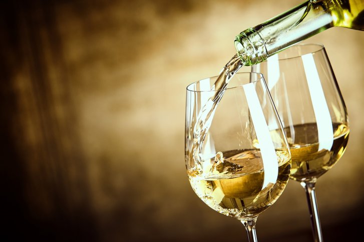 Weiße Flüssigkeit, die in elegante Weingläser eingeschenkt wird, vor einem unscharfen, goldbraunen Hintergrund.