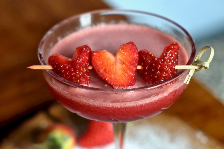 Cocktail im Glas, garniert mit herzförmigen Erdbeeren, auf einem Holztisch serviert.