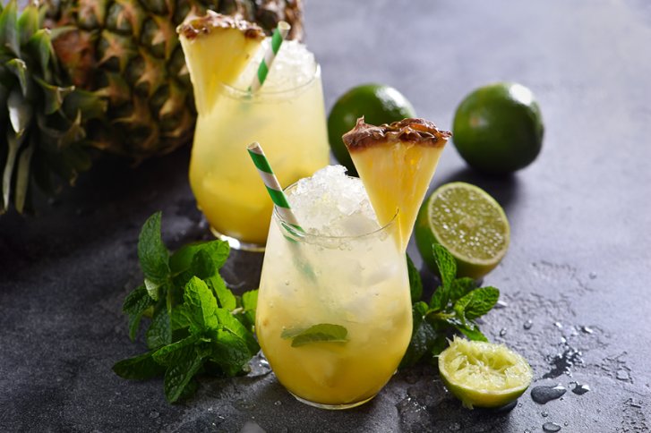 Zwei gelbe Cocktails mit Ananasdekoration und Minze auf einem dunklen, nassen Tisch.