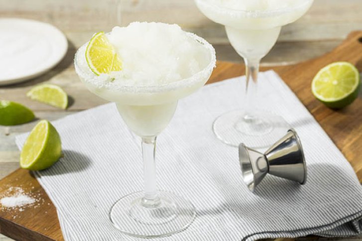 Zwei Margarita-Gläser mit Limettenscheiben, auf Holztisch mit Stoffserviette und Messbecher.