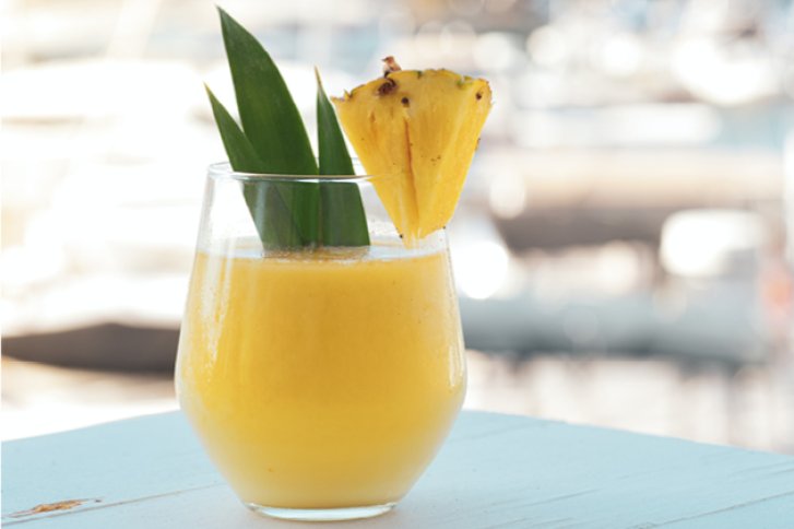 Gelbes Getränk steht auf Holztisch, garniert mit Ananasstück und Blättern, unscharfer Hintergrund.