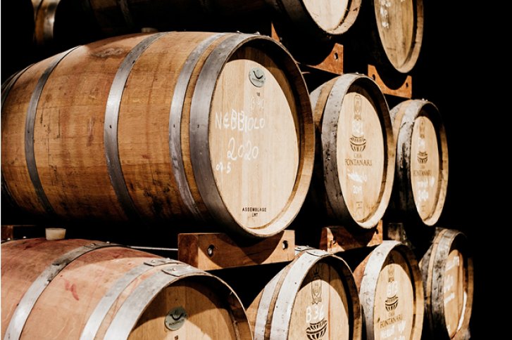 Holzfässer stapeln sich in einem dunklen Lagerraum, mit Aufschrift "Nebbiolo 2020" sichtbar.
