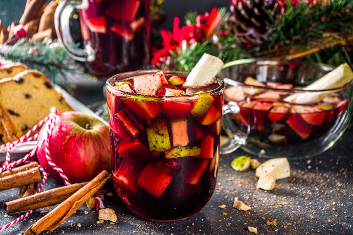 Ein Glas Sangria mit Obststücken steht weihnachtlich dekoriert auf einem Tisch mit Zimt.