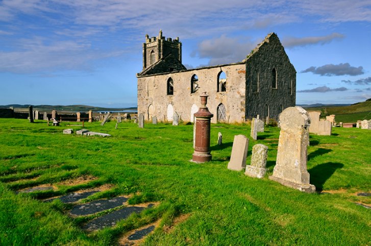 Eine alte, verfallene Kirche steht auf einem Friedhof, umgeben von grünen Wiesen.