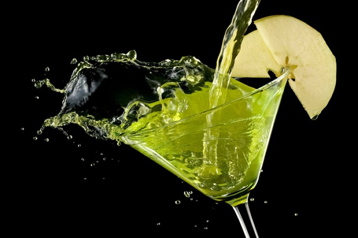 Ein Martini-Glas, gefüllt mit grünem Getränk, daneben schwimmt Apfelscheibe, vor schwarzem Hintergrund.