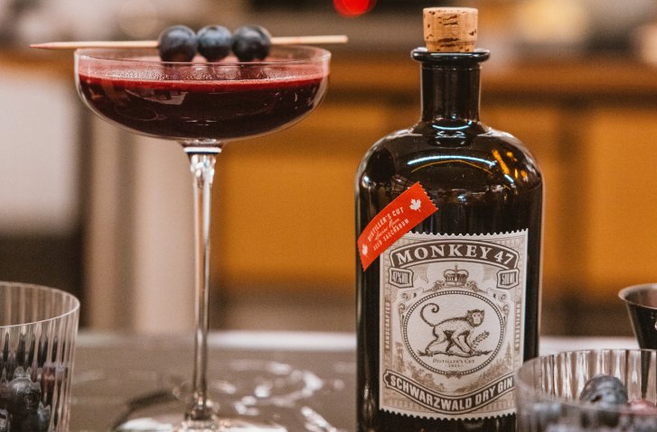 Cocktailglas mit roten Getränk und Heidelbeeren, neben Monkey 47 Ginflasche, auf Tresen stehend.
