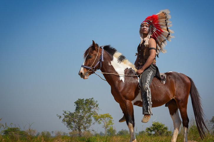 Reiter mit Federkopfschmuck sitzt auf einem gescheckten Pferd in einer offenen Landschaft.