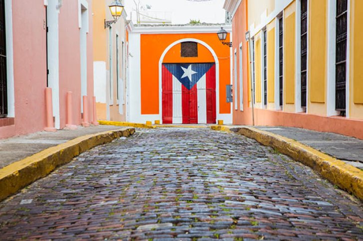Bunte Häuser umgeben eine gepflasterte Straße; Wand mit gemalter Flagge am Ende.