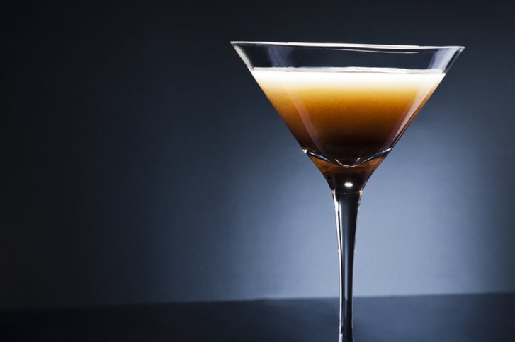 Cocktailglas steht auf dunklem Tisch, gefüllt mit zweifarbiger Flüssigkeit, vor blau-grauer Wand.