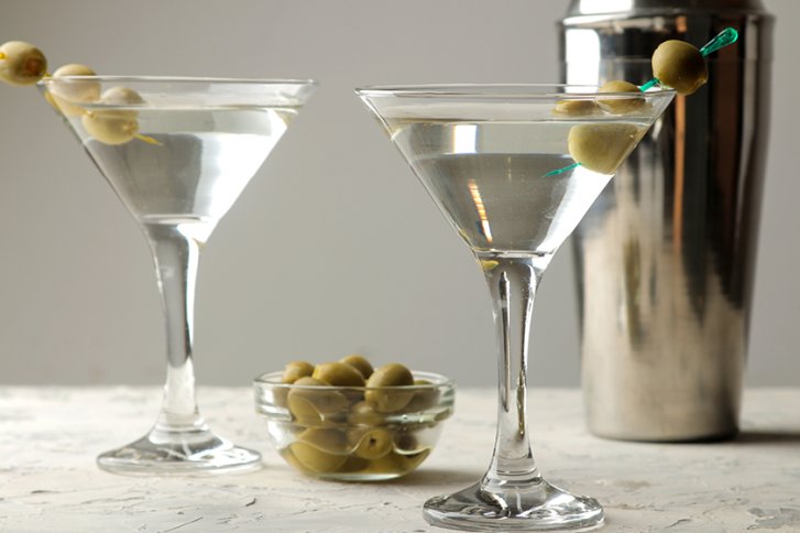 Zwei Martini-Gläser mit Oliven stehen auf einem Tisch neben einem silbernen Cocktail-Shaker.