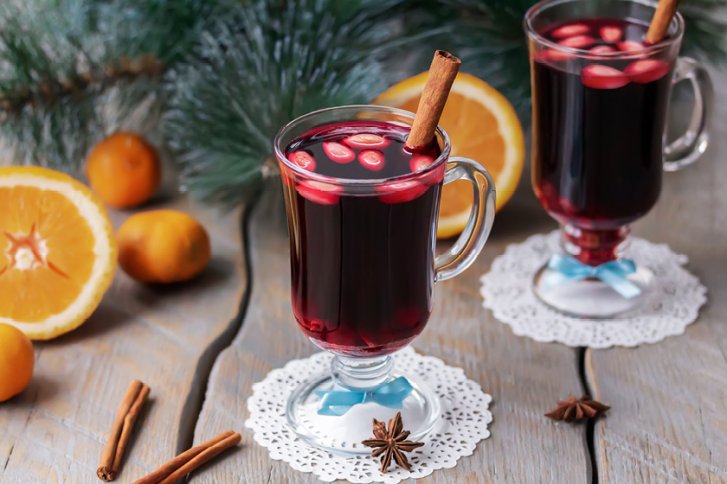 Zwei Gläser Glühwein mit Zimtstangen, auf Spitzendeckchen, umgeben von Orangen und Tannenzweigen.