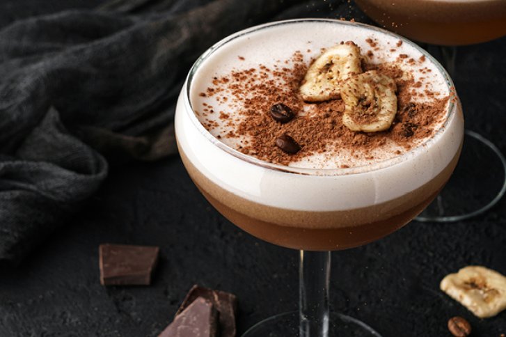 Ein Cocktail mit Schaumhaube steht auf dunklem Hintergrund, garniert mit Bananenscheiben und Kakao.