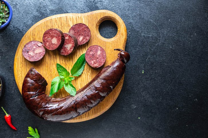 Eine Blutwurst liegt geschnitten auf einem Holzbrett, dekoriert mit frischen Kräuterblättern.