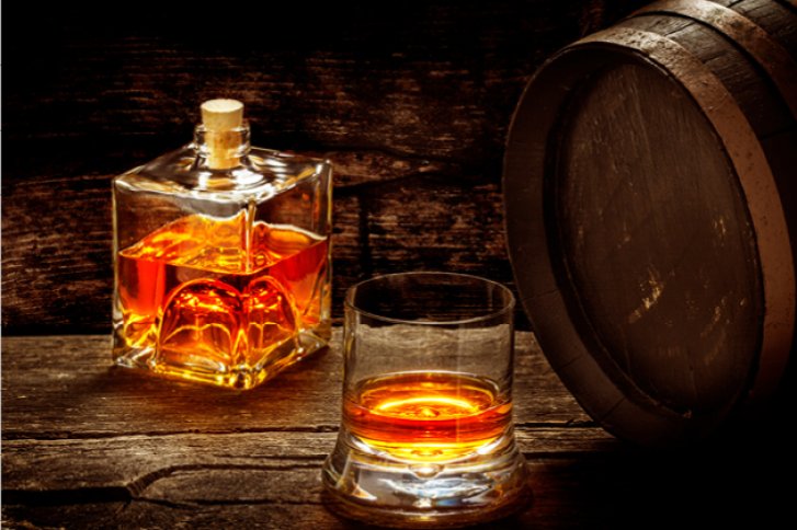 Eine Whiskyflasche und ein Glas stehen auf einem Holztisch neben einem Holzfass.