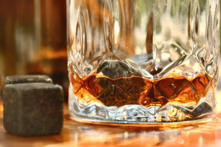 Kristallglas enthält Whisky, daneben sind zwei dunkle Eiswürfel auf einem Holztisch positioniert.