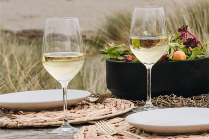 Zwei Weingläser auf einem Tisch, neben weißen Tellern und Salatschüssel im Freien.