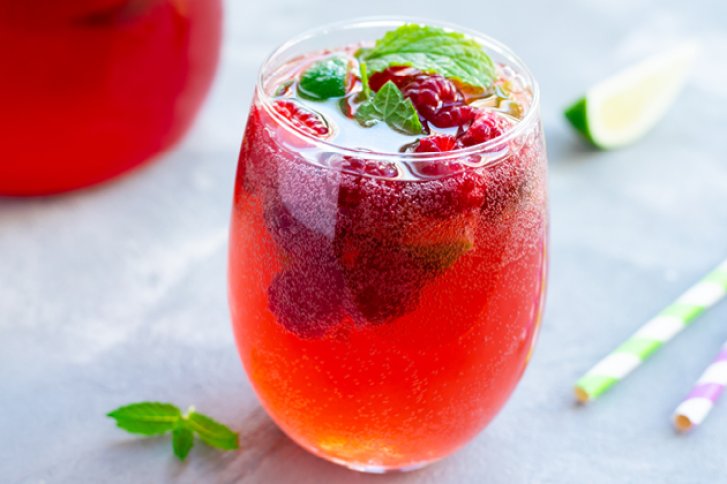 Ein mit Himbeeren und Minzblättern garnierter roter Cocktail steht auf einem grauen Tisch.