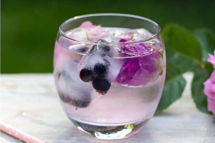Ein rundes Glas enthält rosa Getränk mit Eiswürfeln, Beeren und Rosenblüten auf Holztisch.